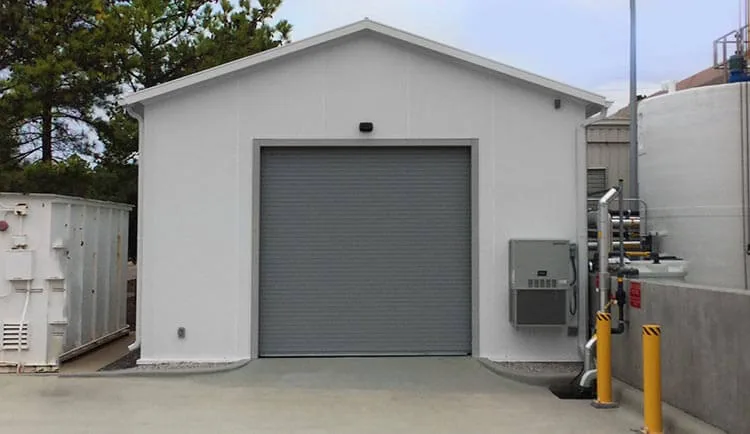 White prefabricated storage building with a roll-up door, wall-mounted HVAC unit and yellow bollards beside process tanks at an industrial plant