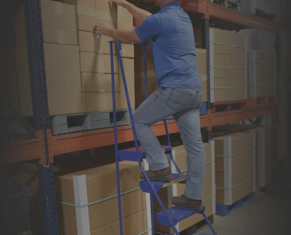 Worker on a blue steel rolling warehouse ladder with handrails reaching cartons on pallet rack