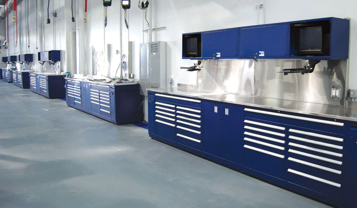 Row of blue service-bay workstations with drawer cabinets, stainless backsplash and overhead monitors along a dealership shop wall