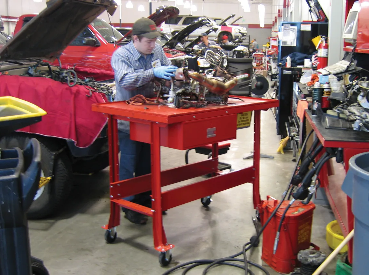 Technician disassembling an engine on a red mobile tear-down bench with drip-edge top and drain in an automotive service shop
