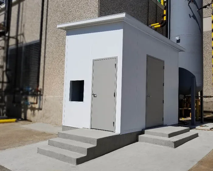 Small white modular storage building with two steel doors and concrete steps, set against an industrial facility wall
