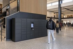 Dark gray smart parcel locker bank with a touchscreen kiosk in a busy university building lobby as students walk past