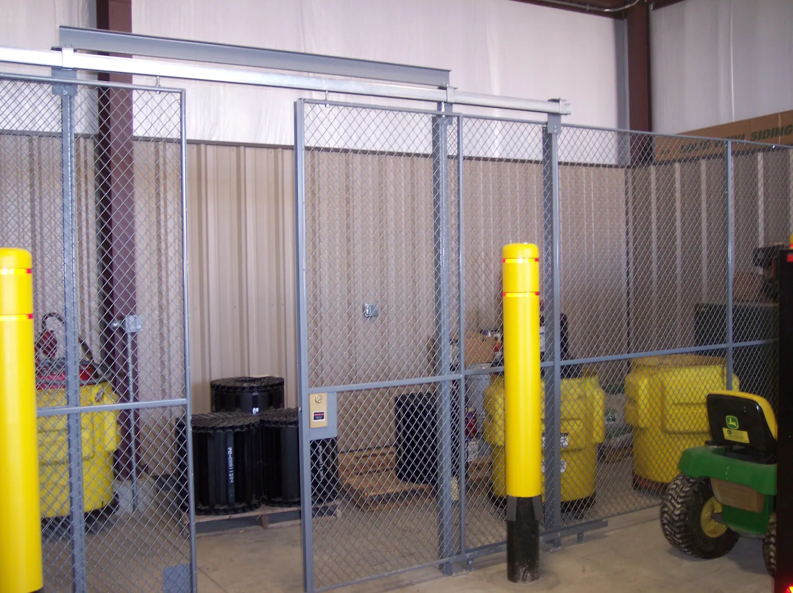 Gray SpaceGuard FordLogan woven diamond-mesh equipment cage with double sliding doors and yellow bollards
