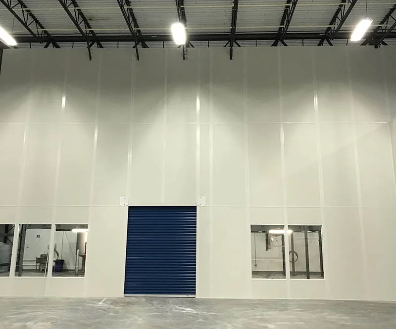 Tall white prefabricated modular divider wall in a warehouse with a blue roll-up door and two banks of windows, reaching up toward the roof joists