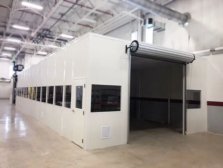 White modular room with windows, a personnel door, a wall vent and an open roll-up door, built inside a manufacturing plant