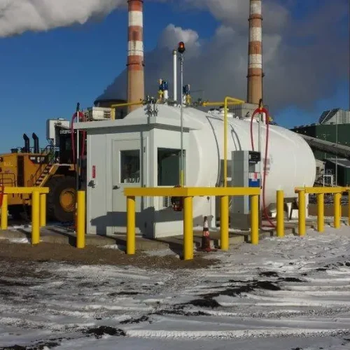 A small industrial building surrounded by yellow safety barriers stands in the foreground, with a large machine adjacent. Nearby, Utah guard booths add a sense of security. In the background, smokestacks emit steam against a clear blue sky. Snow blankets the ground.