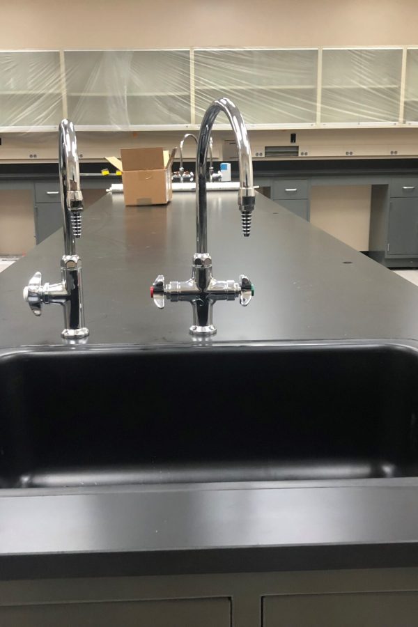 A modern laboratory countertop with two metallic faucets and a black sink basin at Weber State University. The background features cabinets, some with glass doors, and shelves covered with plastic sheeting, giving the room a clean, organized appearance.