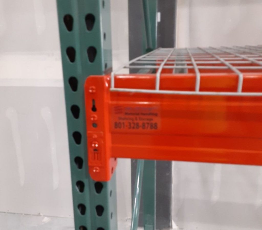 A close-up of an industrial storage rack with a green vertical support beam and an orange horizontal shelf. The visible contact number in black font on the orange part signifies efficient pallet racking in Salt Lake. The background is out of focus.