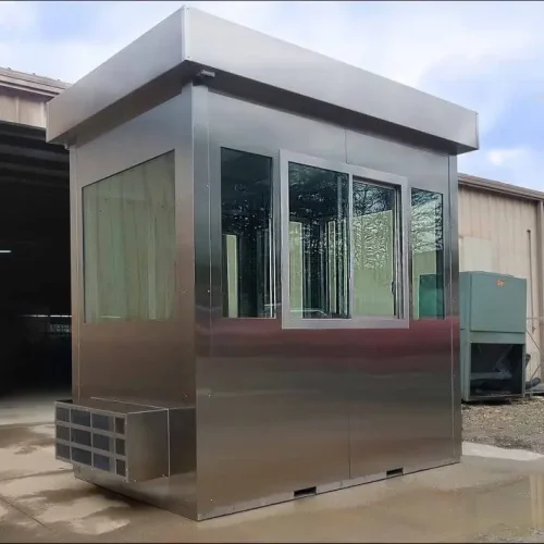 A metallic Utah guard booth with large glass windows on all sides is stationed outside a building. Featuring a flat roof, it rests on a concrete surface, framed by a backdrop of industrial equipment and surrounding architecture.