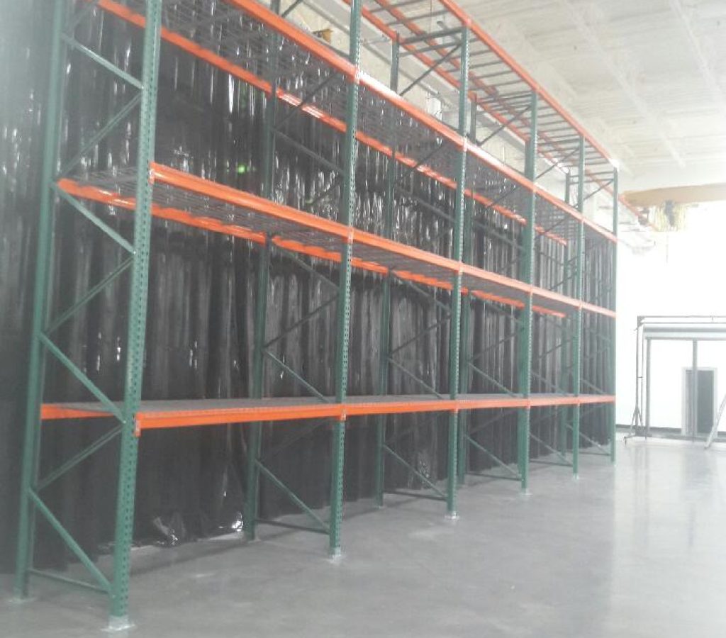 An empty industrial warehouse with tall, metal storage racks stands ready for retrofitting seismic foot plates. The racks are painted green with orange shelves against a wall covered in black material. Gray concrete floors complement the high ceiling and exposed beams, adhering to Utah pallet rack seismic standards.