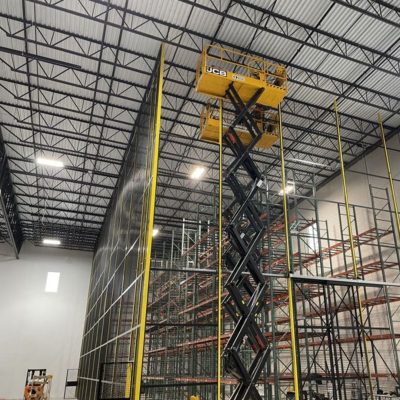 A yellow scissor lift with two workers is elevated near the ceiling of a large warehouse, working on installing or inspecting tall metal shelving units and a safety net along the racks.